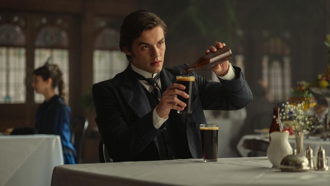 Louis Partridge pouring dark beer from a bottle into a glass.