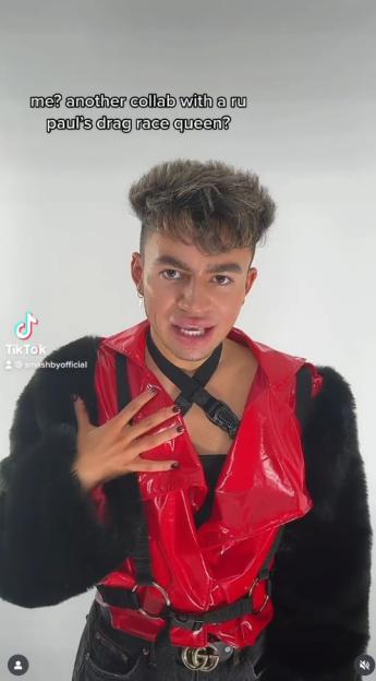 A person wearing a shiny red top with black furry sleeves, and a black choker, with text reading, "me? another collab with a ru paul's drag race queen?"