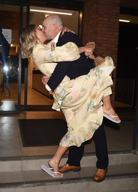 Joe Duttine and Sally Carman sharing a kiss, with Sally holding her sandals and wearing a floral dress, and Joe in a dark suit with tan shoes.