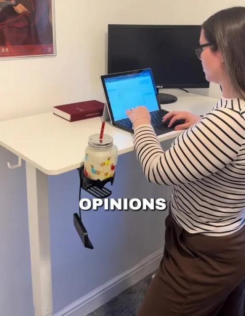 A person sitting at a desk with a laptop, a closed book, a monitor, and a mug on a cup holder that reads "OPINIONS".