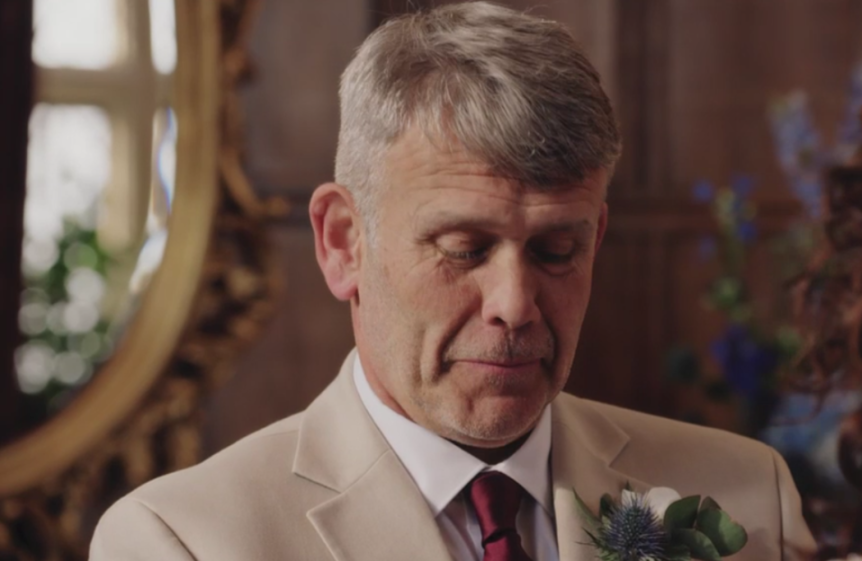 Man in a suit with a boutonnière, looking down with a somber expression.
