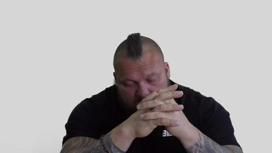 A large man with a mohawk sits with his hands clasped, looking down.