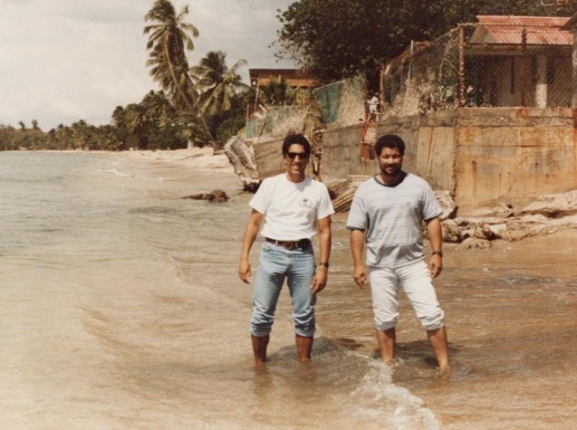 Two men standing in the ocean on a beach with palm trees and buildings in the background.