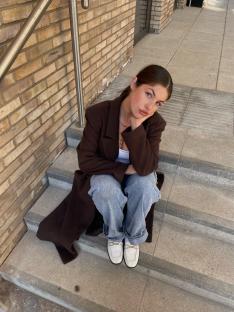 Woman sitting on steps wearing a brown coat and light blue jeans.