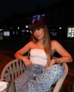 Woman sitting outdoors at night wearing a white top and sequined jeans.