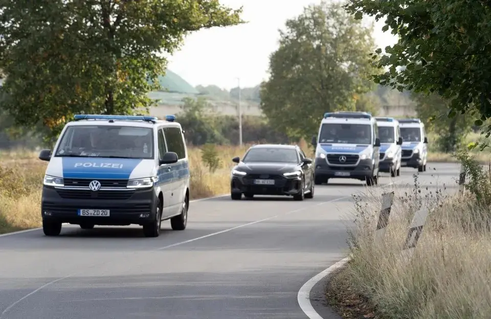 A motorcade of several police vans and a black car on a road.