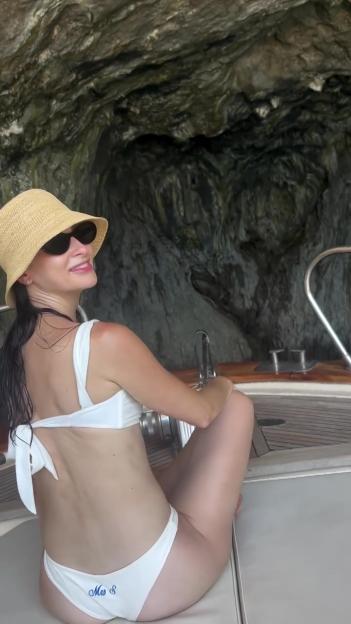 Woman in a white bikini sitting on a boat, looking back at a cave.