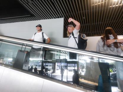 Two men with backpacks ride an escalator at an airport.