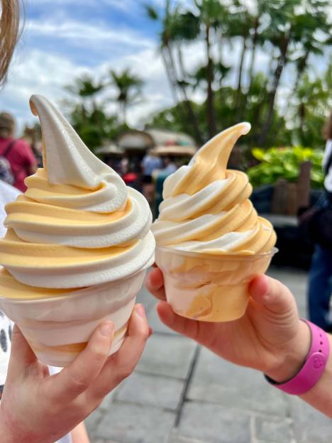 Two hands holding cups of Dole Whip with a blurred background of trees and people.