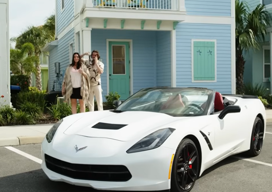 White Corvette convertible parked in front of a light blue house; a man and woman stand nearby.