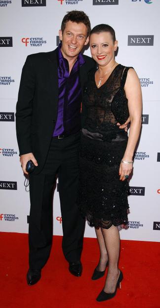A man in a purple shirt and black suit with a woman in a black sequined dress, standing on a red carpet.