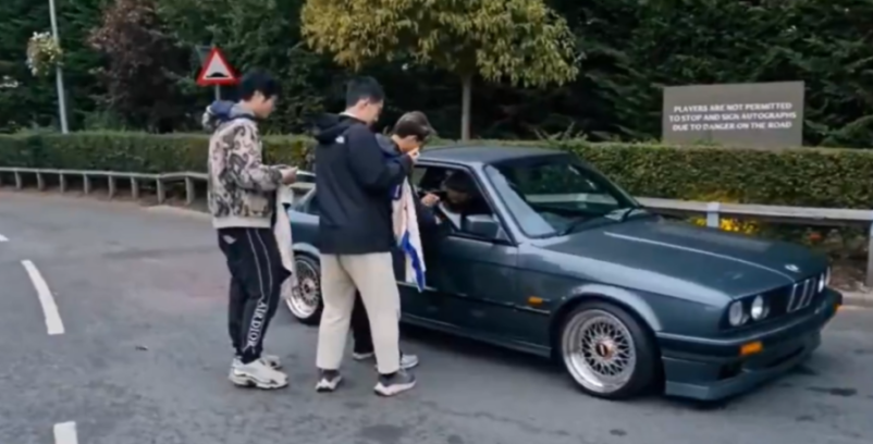 Two men interacting with a driver in a parked car next to a sign that reads: "PLAYERS ARE NOT PERMITTED TO STOP AND SIGN AUTOGRAPHS DUE TO DANGER ON THE ROAD."