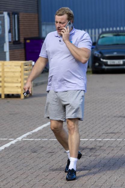 Tom Skinner on a phone call wearing a wedding ring, a light purple polo shirt, grey shorts, and black dance shoes.