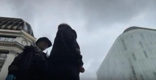 Two people in dark clothing walking in front of buildings.