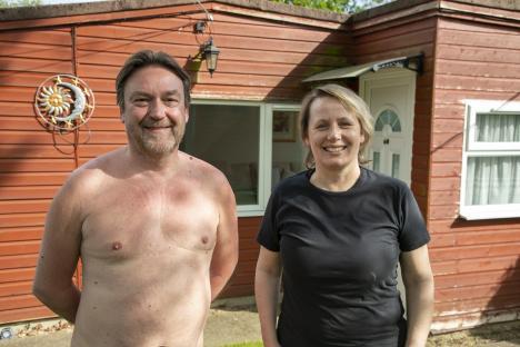 A shirtless man and a woman wearing a black t-shirt stand outside a red cabin.