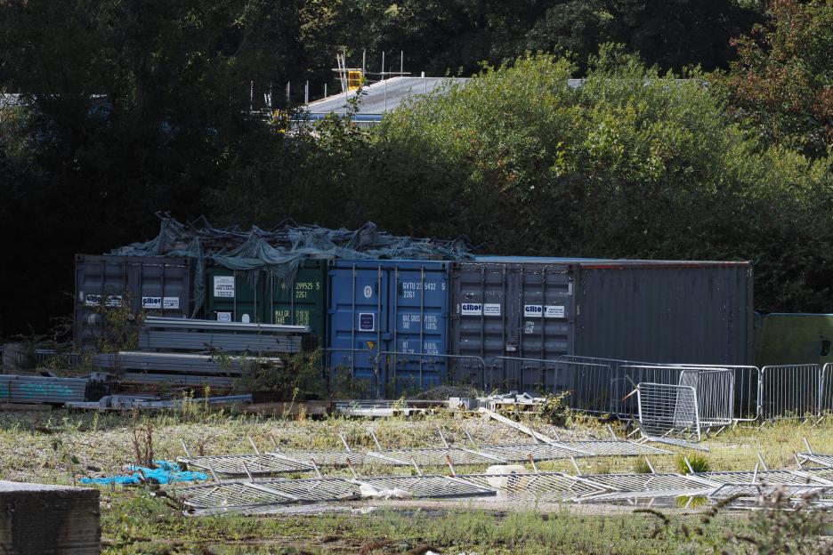 Shipping containers and metal barriers on overgrown ground with trees and scaffolding in the background.