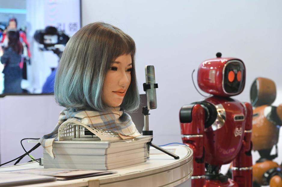 A humanoid robot with blue hair and a scarf, alongside a red robot at a Hong Kong Electronics Fair.