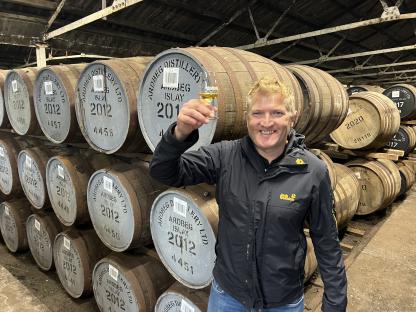 Man toasting with whisky in a distillery surrounded by barrels.