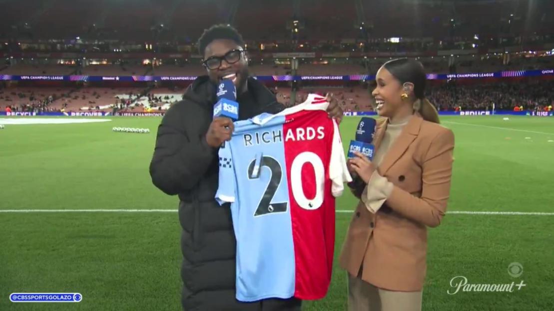 Micah Richards holds up a half red, half light blue soccer jersey with "RICHARDS 20" on the back, next to a smiling woman on a soccer field.