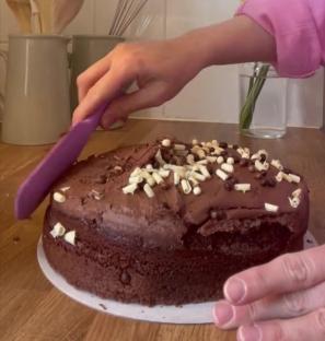 A person cutting into a chocolate cake decorated with white chocolate shavings.
