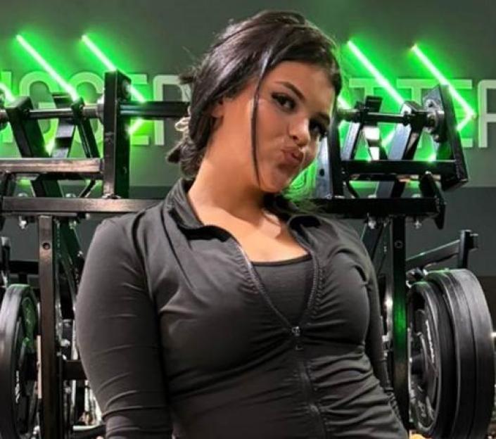 Woman in black zip-up top posing in front of gym equipment.