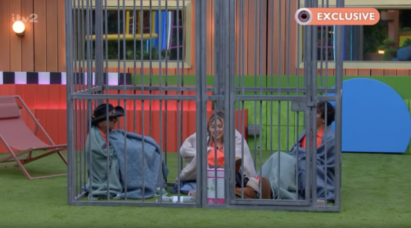 Three people sit inside two adjacent metal cages on a green floor, with an "EXCLUSIVE" banner in the top right.