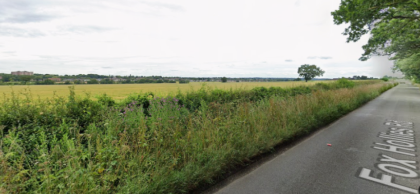 A paved road with "Fox Hollies Rd" painted on it, running alongside a field with a town in the distance.