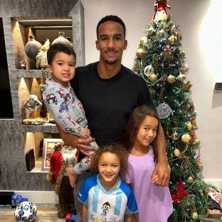 A man holding two young children in front of a decorated Christmas tree, with another young child standing beside him.