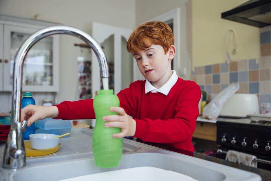 A young boy in a red school uniform fills a green water bottle from a kitchen faucet.