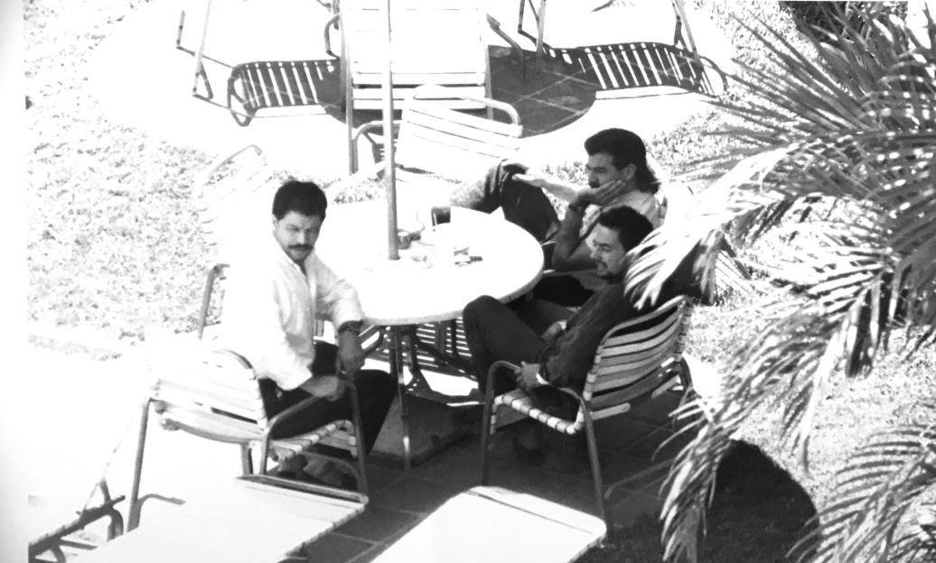 Black and white photo of three men sitting around a table with drinks outdoors.