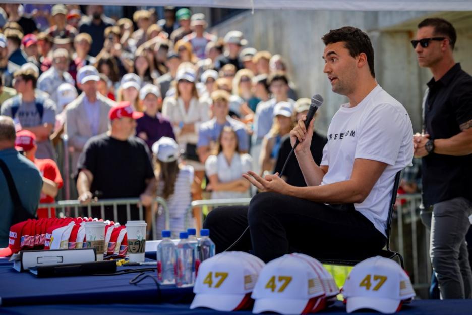 Man speaking into a microphone at an outdoor event with a crowd in the background.