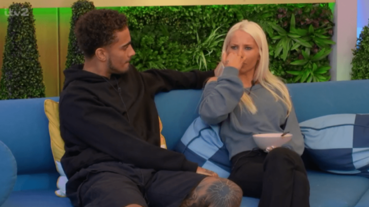 A man with dark curly hair comforting a blonde woman who is wiping her nose while holding a bowl.