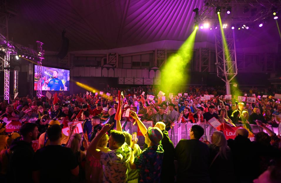 Spectators at a darts match watch a large screen displaying Luke Littler.