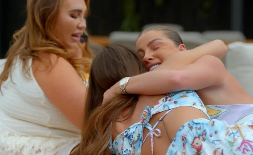 A woman with light brown hair and a white top looking concerned, watching two other women hugging, with the woman on the right crying.