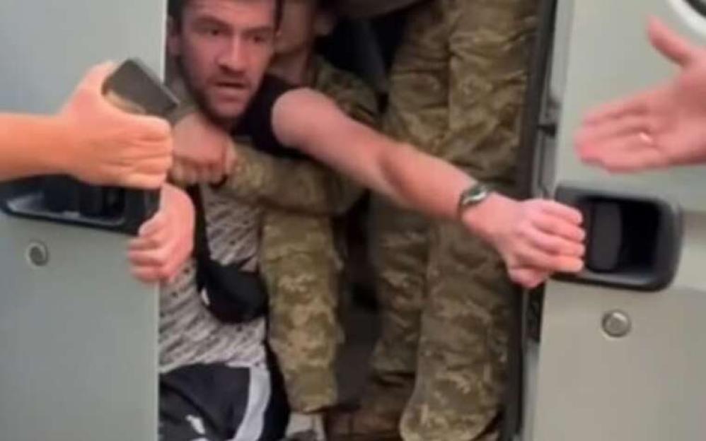 Man being held by two people in camouflage entering a vehicle while another person tries to pull him out.