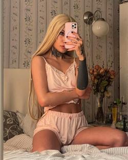 Woman with long blonde hair and tattoos taking a selfie in pink pajamas.