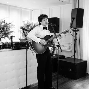 Black and white photo of a man singing and playing an acoustic guitar at a party.