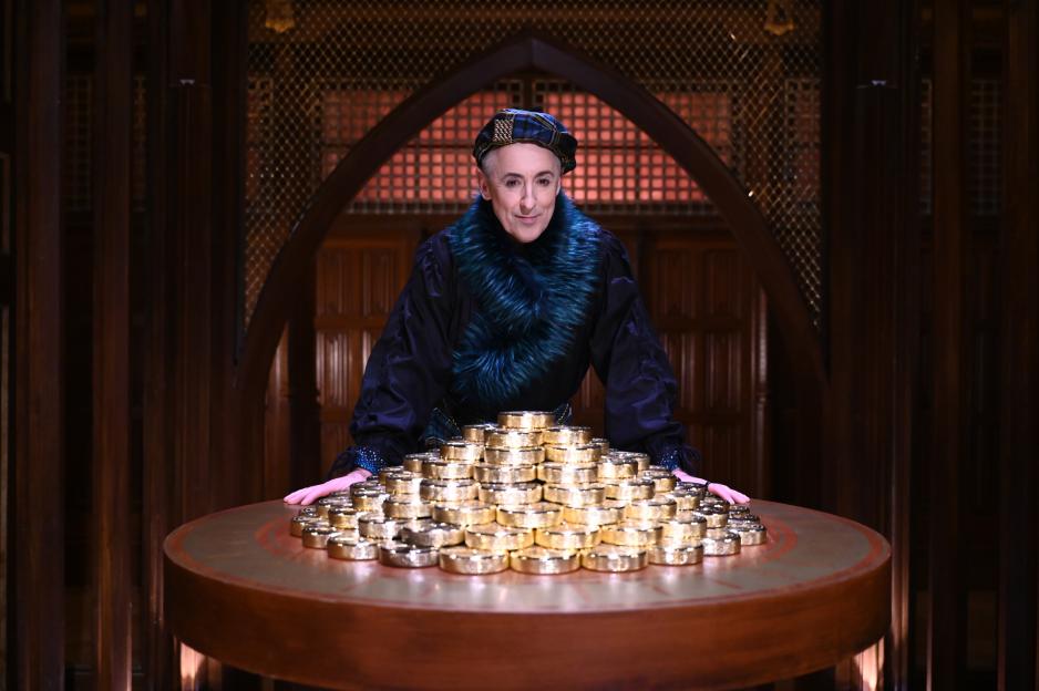 Alan Cumming leaning on a table with a large pile of gold coins.