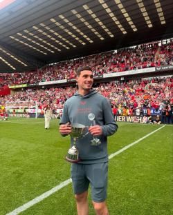 Footballer Lloyd Jones holding two trophies on a football pitch.