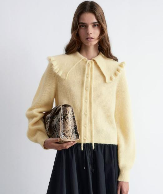 Model wearing a ruffled collar cardigan in light yellow, a black skirt, and holding a snakeskin handbag.
