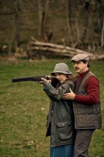 A woman aims a shotgun while a man assists her.