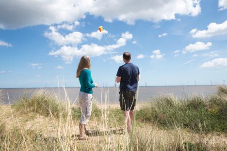 Couple flying a kite on a beach with wind turbines in the distance.