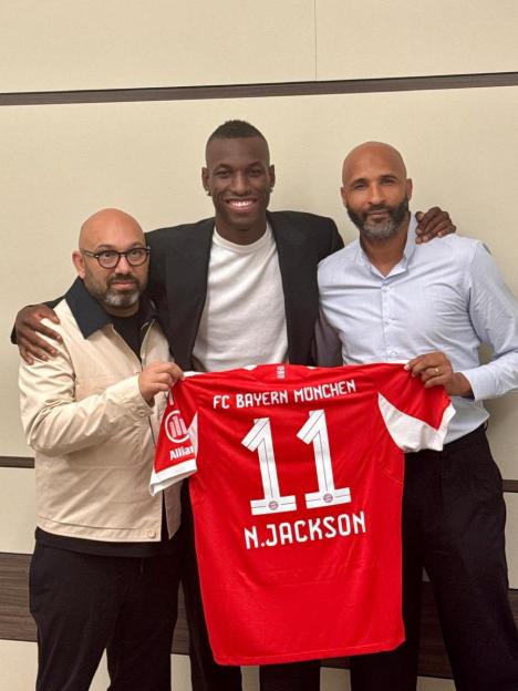 Three men holding up an FC Bayern München jersey with the number 11 and N. Jackson on the back.