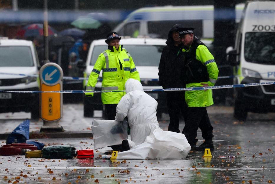 A forensic team member in a white suit works on the ground at a crime scene while two police officers in high-visibility jackets stand nearby, all under falling rain.