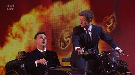 Screenshot of two men in suits on a vintage motorcycle and sidecar.
