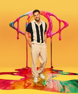 Chris Hughes standing in front of a large, colorful eye graphic for Celebrity Big Brother.