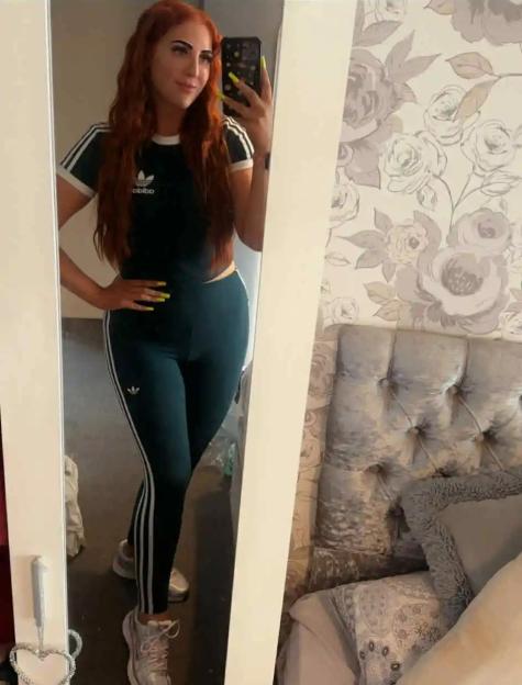 A woman with long red hair wearing a dark green Adidas crop top and leggings takes a mirror selfie.