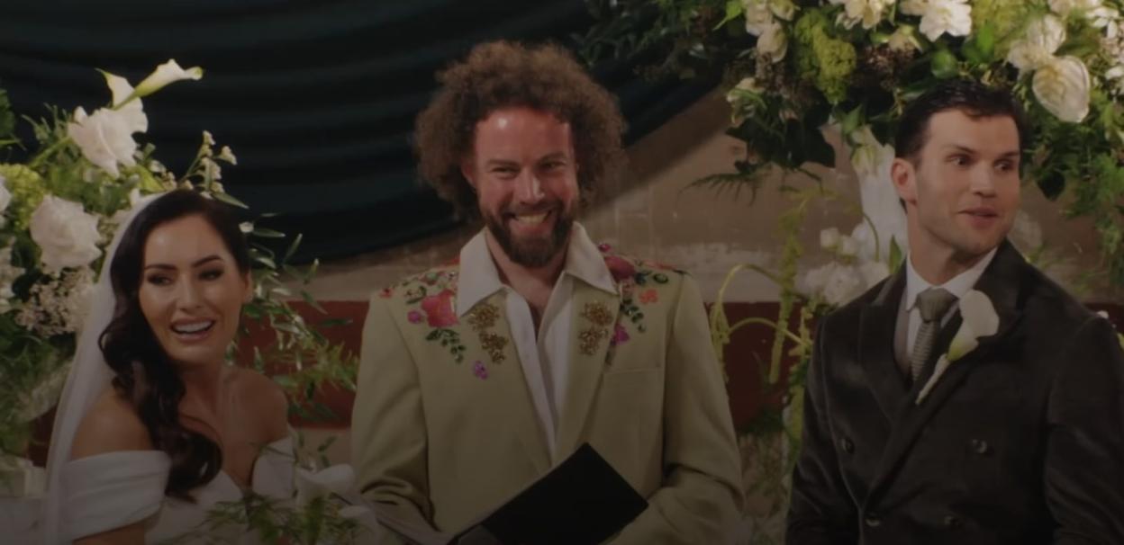 A bride smiling, a man with an afro smiling, and a groom glancing aside during a wedding ceremony.