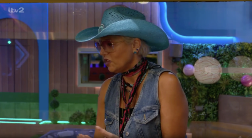 A woman with a short blonde hair wearing a blue cowboy hat, pink sunglasses, a denim vest, and a patterned scarf is speaking.