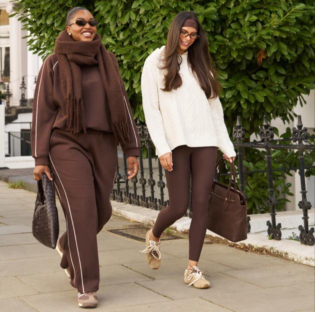 Two women walking down a city street, one in a brown tracksuit and scarf, the other in a white sweater and brown leggings.